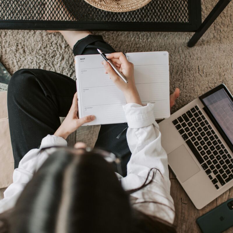 Top view of anonymous woman in casual wear sitting on floor with laptop and smartphone and creating plan on notebook while resting during break in modern living room